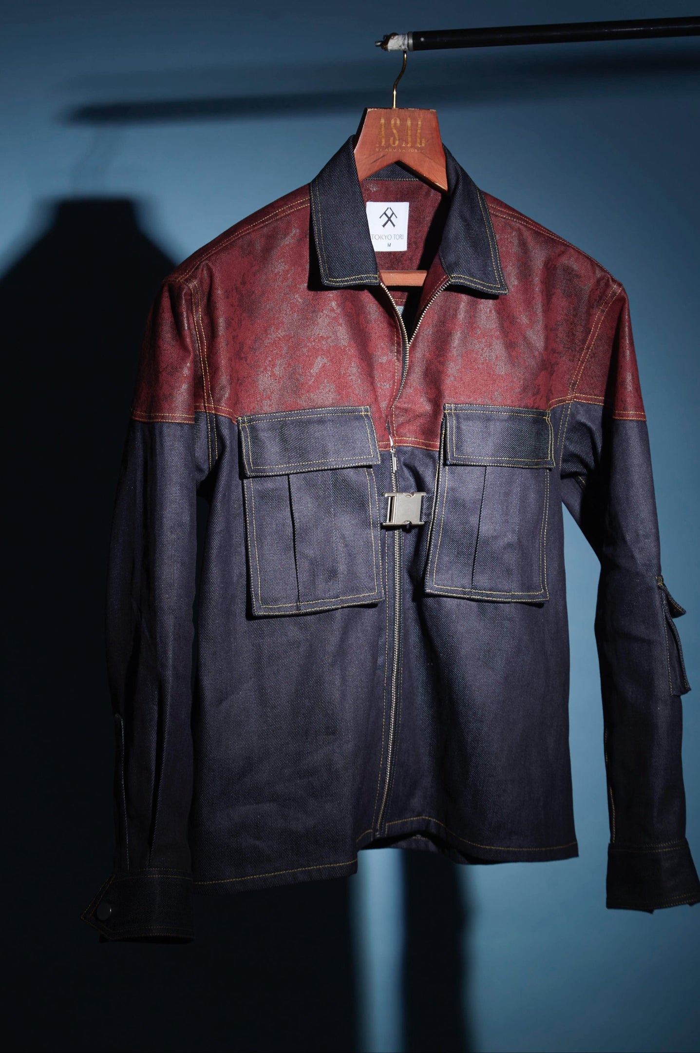 A two-tone leather jacket hanging on a wooden hanger against a dark blue-gray background. The upper chest and shoulders are a distressed reddish-brown leather, while the sleeves, collar, and lower body are a dark navy blue leather.